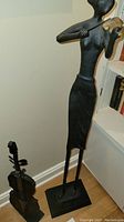 Full view of the black metal woman playing violin sculpture next to the smaller violin sculpture on stands on the floor