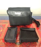 Three black carrying cases of different sizes for the binoculars, showing brand name 'Bushnell' on the largest case.