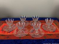 Six crystal dessert cups displayed on six matching plates arranged on a decorative cloth. Each set has a pinwheel crystal cut pattern.