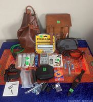 Overview of lot contents on a blue cloth including leather bags, travel bottles, toiletry items, razor, alarm device, luggage scale, adapters, and more.