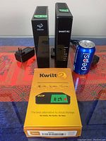 Kwilt 2 in box with Hitron and Smart RG modems and power adapters beside a Pepsi can for size reference