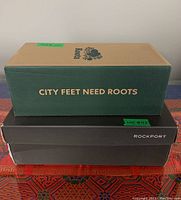 Three stacked shoe boxes: Roots box on top with slogan 'City Feet Need Roots', middle Rockport box, and bottom Rockport box.
