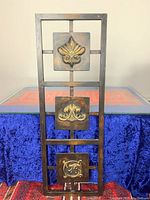 Front view of the first metal outdoor decor panel featuring three ornate square plaques with symmetrical floral designs in distressed black, bronze, and gold finish.