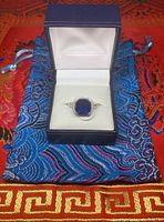 Blue sapphire ring in sterling silver with halo of cubic zirconias, displayed in navy blue box with white velvet interior and blue fabric pouch.