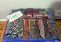 Nine men's ties laid out side by side showing various patterns and colors, including boxed packaging above them