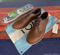 Pair of men's brown leather TOMS shoes size 8/40 displayed on blue and red patterned surface with blue and white TOMS branded drawstring shoe bag and sealed Comfort Foot memory insoles next to shoes.