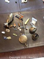 Photo showing multiple small collectibles including metal pins, two salt shakers, clock weight, and a tiny oil lamp part placed on a dark surface.