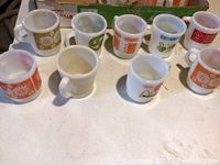 Top view of 10 colorful vintage Fire King milk glass mugs with various patterns and one McDonald's mug showing wear.