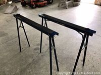 Photo showing two metal sawhorses with rectangular beams and foldable legs in a garage setting.