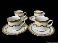 Four porcelain demi-tasse cups with matching saucers arranged in pairs on black surface showing floral and gold trim design.