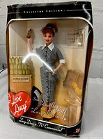 Front view of Mattel I Love Lucy collector edition doll in packaging, showing Lucy Ricardo dressed in gray checkered outfit holding a VitameATUAEGAMIN bottle with advertising props.