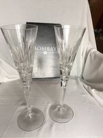 Two clear crystal champagne flutes with vertical cut designs and slender stems, standing in front of a Bombay Company box.
