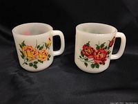 Two vintage milk glass mugs with rose patterns, one showing yellow and red rose designs on each mug's sides, placed side by side