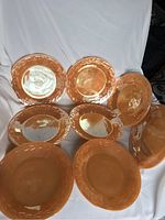Eight Fire King dinner plates with peach lustre glaze showing raised laurel leaf pattern on rims, arranged on a white cloth background. Light reflection highlights wear and surface condition.