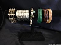 Photo showing a total of seven bracelets on a black velvet display stand: three expanding types with various bead and metal designs on left, four solid glass bangles in green, brown, amber and honey colors on right.