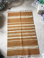 Photo of a brown rectangular throw rug with cream horizontal stripes and fringe on shorter ends, laid out on a carpeted floor.