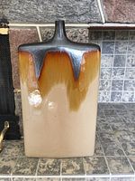 Photo showing front of large rectangular ceramic vase with dark gray to amber glaze at the top, cream base, on tiled surface in front of stone wall.