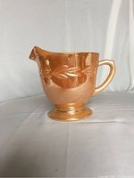 Side profile of vintage Fire King peach lustre creamer highlighting the embossed laurel pattern and handle shape.