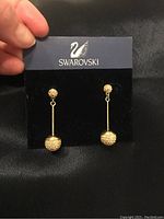 Front view of Swarovski gold-tone dangle earrings on branded card. One earring has a missing backing plate as noted.