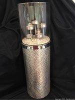 Front view of the tall candle holder showing the textured silver metal base and clear glass upper with seven tealight candles on raised holders inside, resting on white decorative stones.