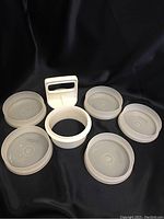 Photo showing five round white mold bases with translucent lids, a white circular ring mold, and a white handle press tool on black fabric background.