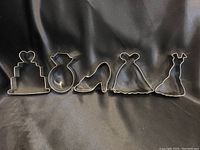 Five metal wedding themed cookie cutters displayed on a black fabric background, including cake, ring, shoe, and two wedding gowns.