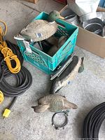 Five plastic mallard duck decoys with green heads and detailed feathers, placed around and inside a green milk crate on a concrete floor. Black cords and metal buckets visible nearby. An anchor line is visible attached to at least one decoy.
