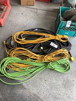 Three bundles of extension cords in yellow, green, and black colors coiled on a concrete floor.