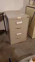Side and front view of beige metal file cabinet showing two drawers with label holders and lock on top drawer.