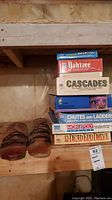 Photo of six board games stacked on a wooden shelf plus three glass items in front