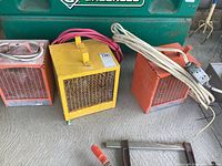 Three industrial-style electric heaters arranged side by side on a concrete floor, showing front grills, cables, and handles