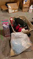 Photo showing plastic bin with assorted holiday stuffed toys including Santa Claus and another red figure, alongside a plastic bag filled with more stuffed toys and a boxed white Easter tree decoration.