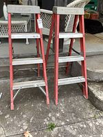 Two red fiberglass framed step ladders with aluminum steps, standing outdoors on concrete ground with wicker furniture in background.