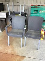 Two grey plastic patio chairs with metal legs standing side by side. Woven pattern visible on seat and back. One chair has armrests, the other does not.