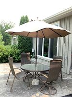 Patio set arranged outdoors showing glass top round table with four brown woven metal chairs and large patio umbrella.