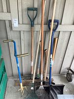 Full view of assorted garden tools including shovels, rakes, hand tiller, and axes showing overview of lot condition and types.