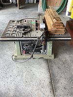 Front view of Delta 10-inch table saw with power cord and stacked wood pieces on top.