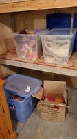 Photo showing three plastic storage bins, one blue and two clear, plus a cardboard box containing various items including toys and fabric pieces.