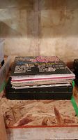 Stack of vinyl LP records on top of black carrying case for cassettes, viewed from side.