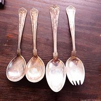Photo of four large silver utensils laid out flat on wooden surface, showing front and side views of spoons and spork