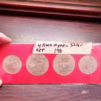 Front side view showing Olympic-themed designs and denominations of four silver coins in red packaging with label.