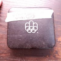 Closed brown vintage case with Olympic rings logo and handwritten label indicating '4 Piece Montreal set silver 203'.