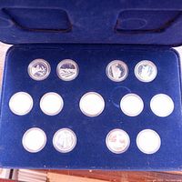 Blue velvet box with 12 silver provincial quarters in protective plastic capsules, arranged in two rows, with some capsules empty.