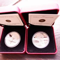 Two silver coins in red display boxes with black interiors, showing detailed engravings and certificate cards.