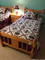 Wood veneer twin bed with carved turned posts on the headboard and footboard, dressed in maroon-green star-patterned quilt and matching pillow, with a bedside lamp and nightstand visible.