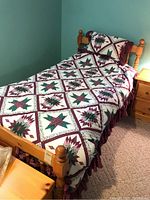 Twin bed made of wood veneer with mattress and burgundy-green cream star-patterned quilt and pillow. Room with teal wall and beige carpet. Small wooden nightstand and lamp present.