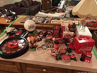 Photo of multiple Coca-Cola collectibles including dolls, mugs, lights, pins, glassware and mini plastic cooler container spread on countertop and table.