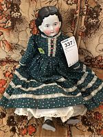 Full front view of antique doll seated on floral couch, showing green dress with lace and buttons, white underskirt, and china head.