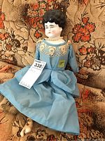 Antique china head doll sitting on fabric with floral pattern, showing full body including leather hands and feet in blue dress with lace collar and pearl necklace.