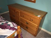 Front view of a wood dresser with 6 drawers showing natural pine finish and mixed metal pulls, placed next to a bed in a room with teal walls